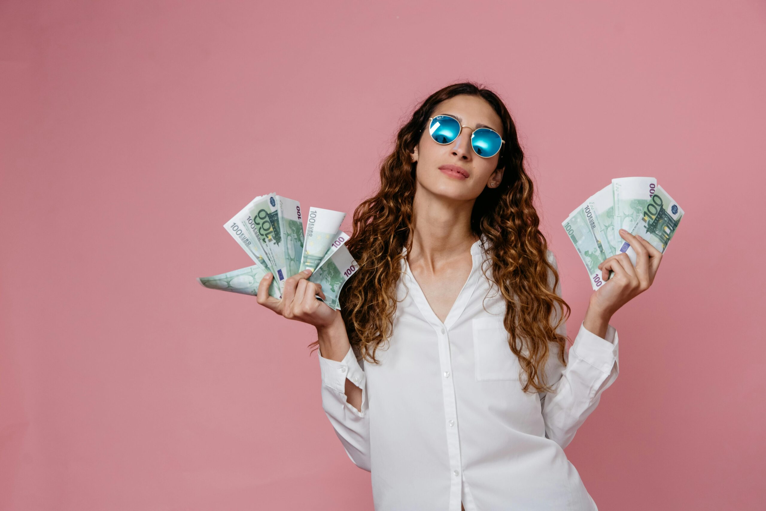 Elegant woman in sunglasses holding euro banknotes against pink background, showcasing confidence and financial empowerment.